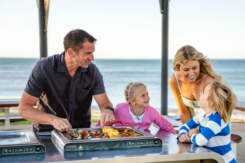 A family of four gathers around an outdoor grill. The man is cooking while the woman and two children, a girl in pink and a boy in stripes, smile and chat. The sea and sky provide a serene backdrop.