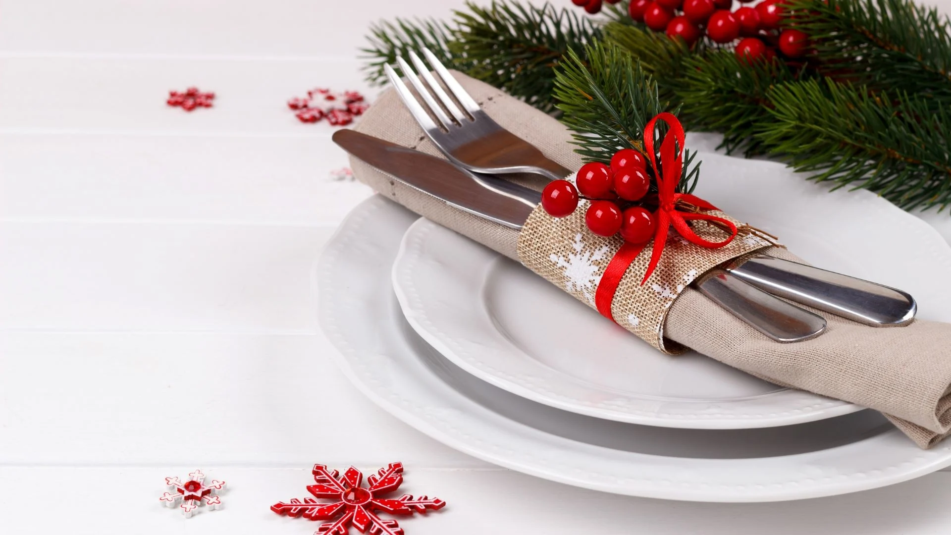 Festive table setting with white plates, a napkin, and cutlery tied with burlap and red berries. Pine branches and red snowflake decorations enhance the white wooden background, creating a holiday-themed arrangement.