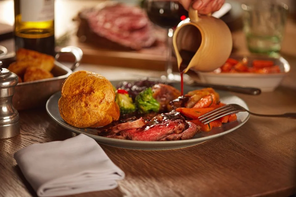 A plate of roasted meat slices, Yorkshire pudding, and assorted vegetables being drizzled with gravy from a jug. A glass of red wine and a napkin are beside the plate on a wooden table.