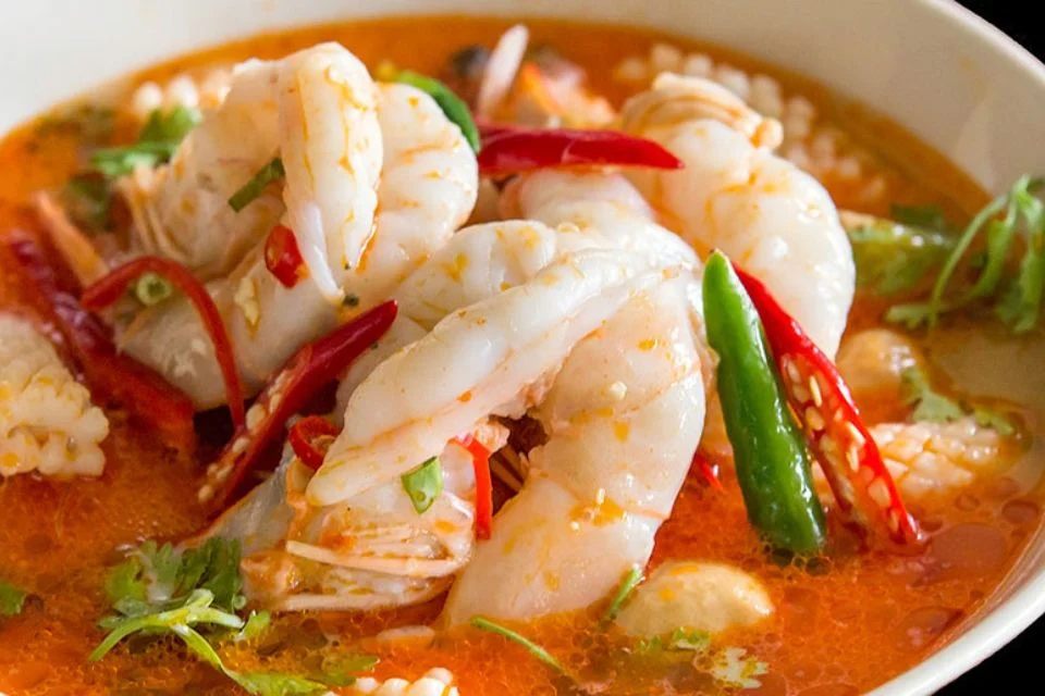 A bowl of spicy shrimp soup garnished with red and green chili peppers, cilantro, and a light broth. The shrimp are plump and pink, surrounded by a vibrant orange broth, creating an appetising and colourful dish.