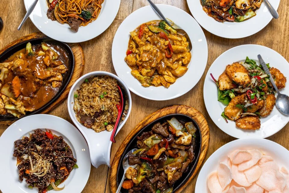 A table filled with various dishes of Chinese cuisine, including stir-fried noodles, fried rice, sizzling beef, vegetables, crispy chicken, and prawn crackers. Each dish is elegantly presented on white plates.