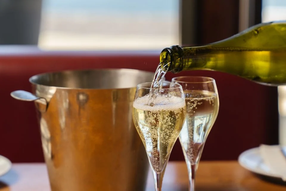 A close-up of two champagne glasses being filled, with a metal ice bucket in the background. The scene is set on a wooden table, with a blurred view through a window suggesting an outdoor or seaside setting.