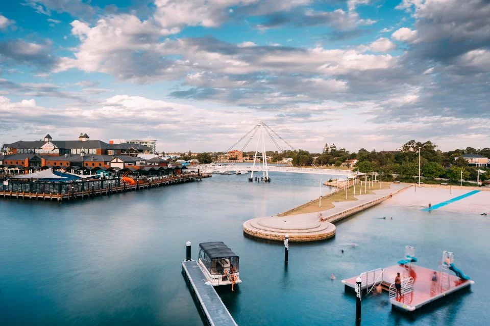 A scenic waterfront with a marina, featuring several boats docked along piers. A large pedestrian bridge is in the background. Buildings with dark roofs line the left side under a partly cloudy sky, and a few people are near the water.