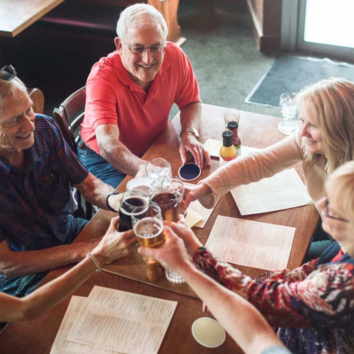 A group of older adults sitting around a table in a restaurant, cheerfully clinking glasses together in a toast, with menus and drinks on the table.