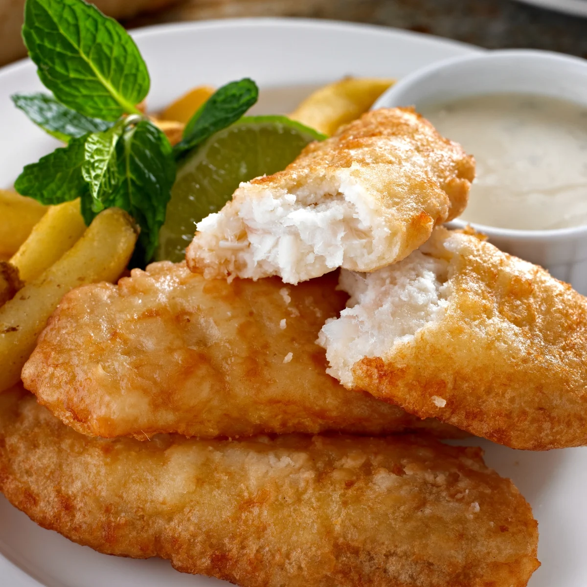 Close-up of golden-brown fish fillets, some broken to show the white flaky inside, on a plate with thick-cut fries. Garnished with fresh mint and a lime slice, accompanied by a small bowl of creamy dipping sauce.