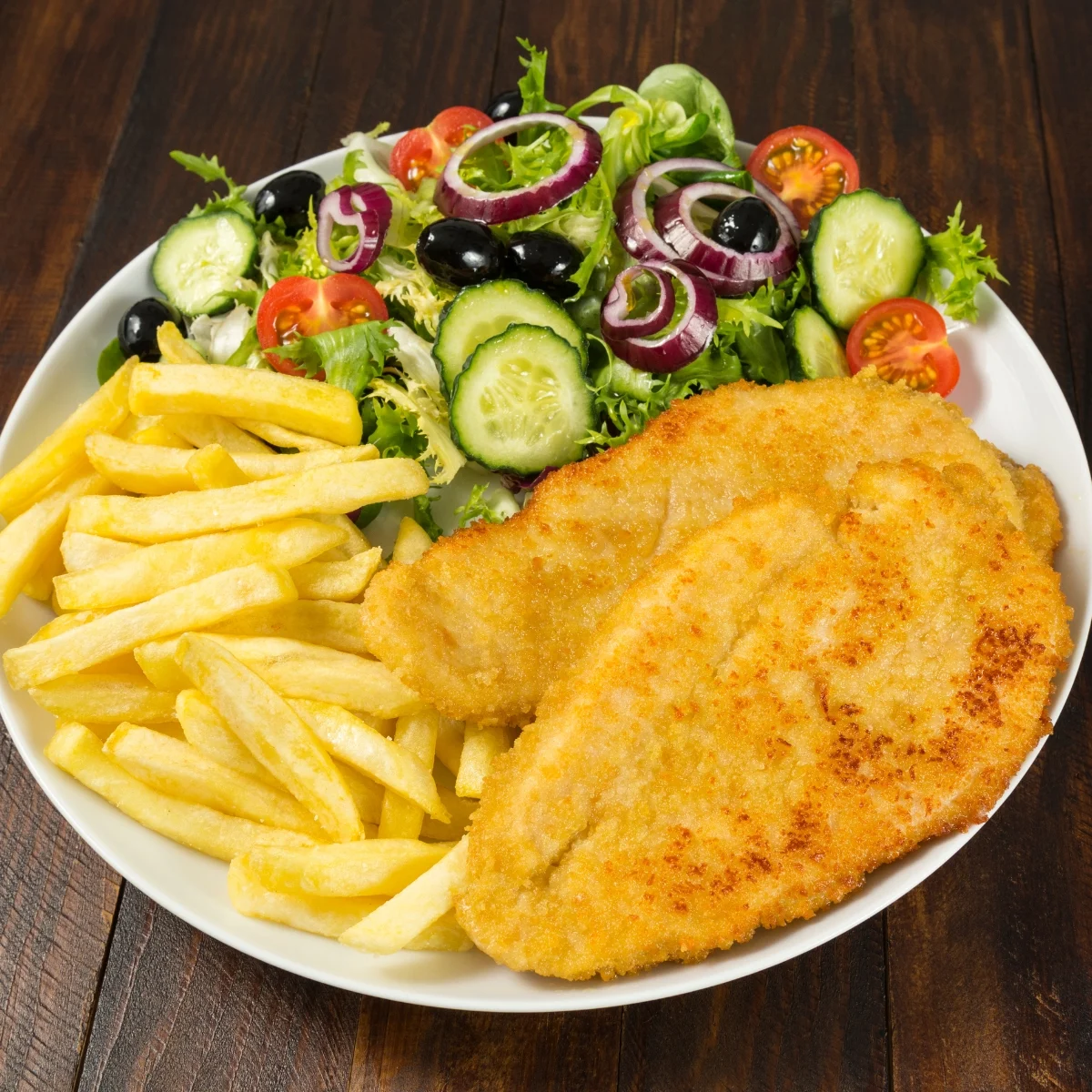 A plate of crispy breaded schnitzel with golden French fries and a fresh salad of lettuce, cucumbers, cherry tomatoes, red onions, and black olives on a wooden table.
