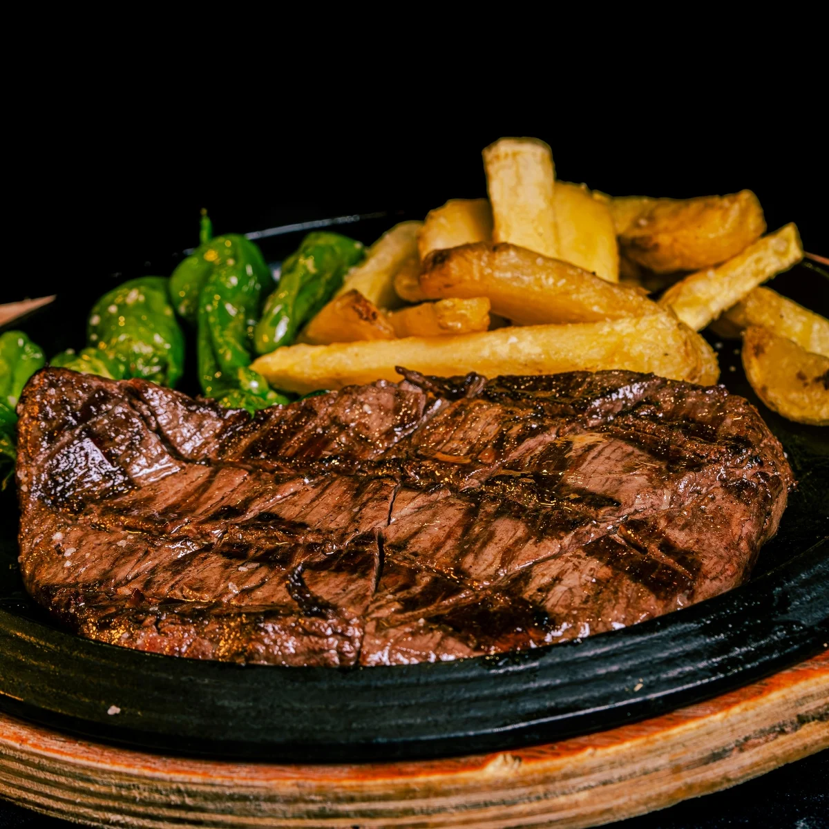Grilled steak with crisscross grill marks on a black plate, accompanied by green peppers and golden-brown French fries, set against a dark background.
