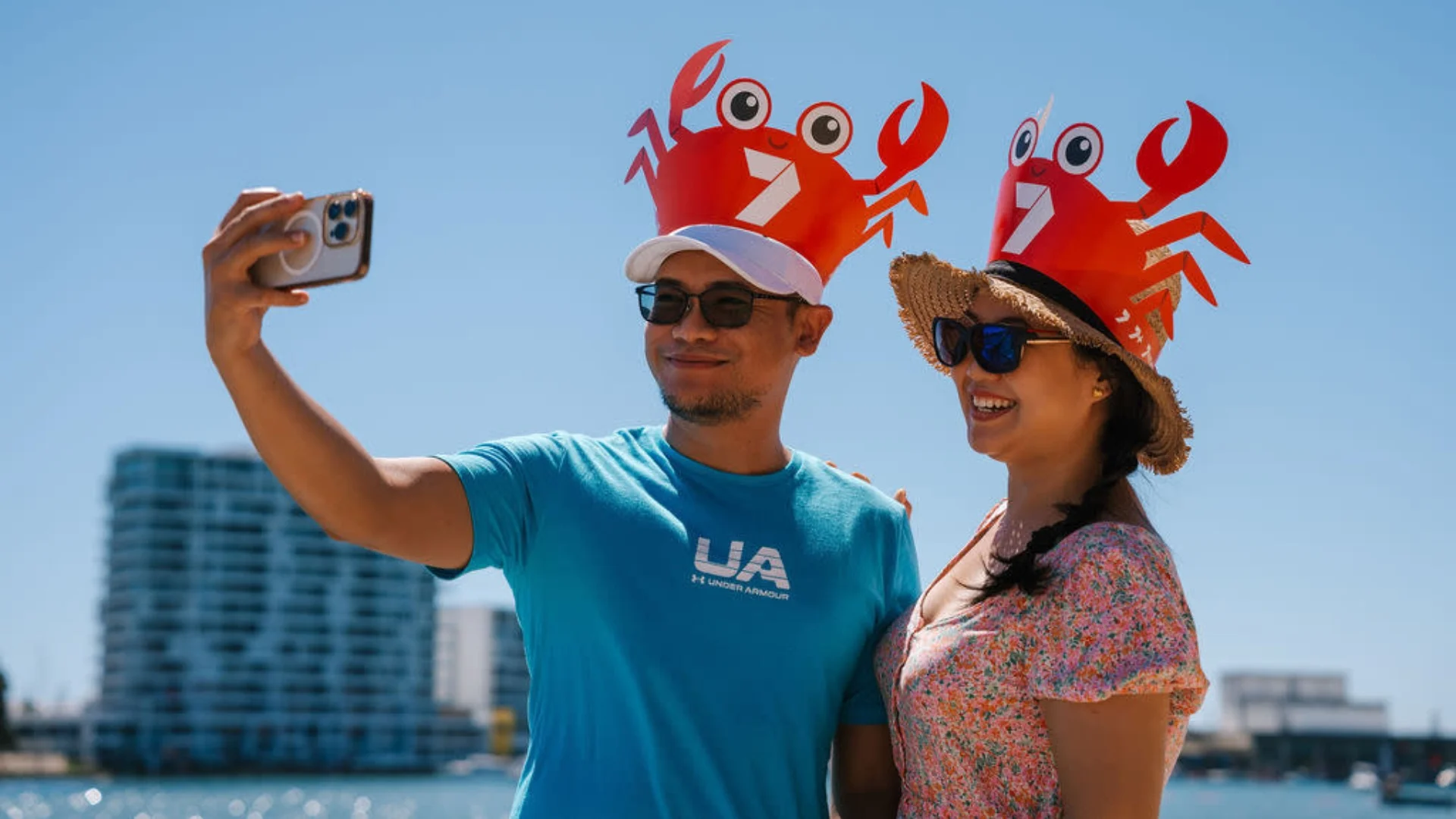 A man and woman smile for a selfie, both wearing crab-themed hats with large eyes and claws. The man is in a blue shirt and sunglasses, while the woman wears a floral dress and straw hat. They stand by a waterfront with modern buildings in the background.