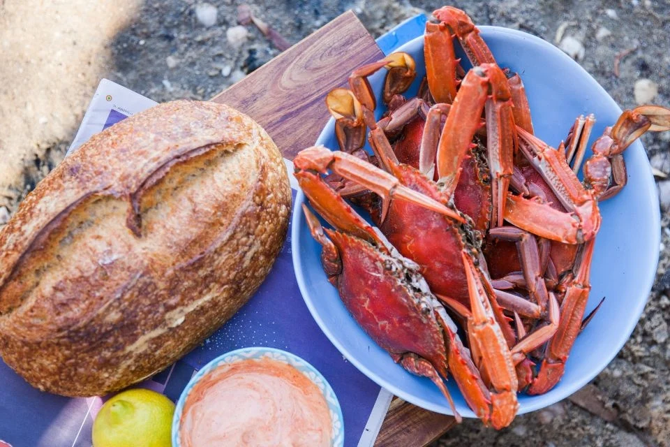 A crusty loaf of bread sits beside a blue bowl filled with steamed red crabs. A small dish of dipping sauce and a lemon are partially visible, all placed on top of a wooden cutting board outdoors.