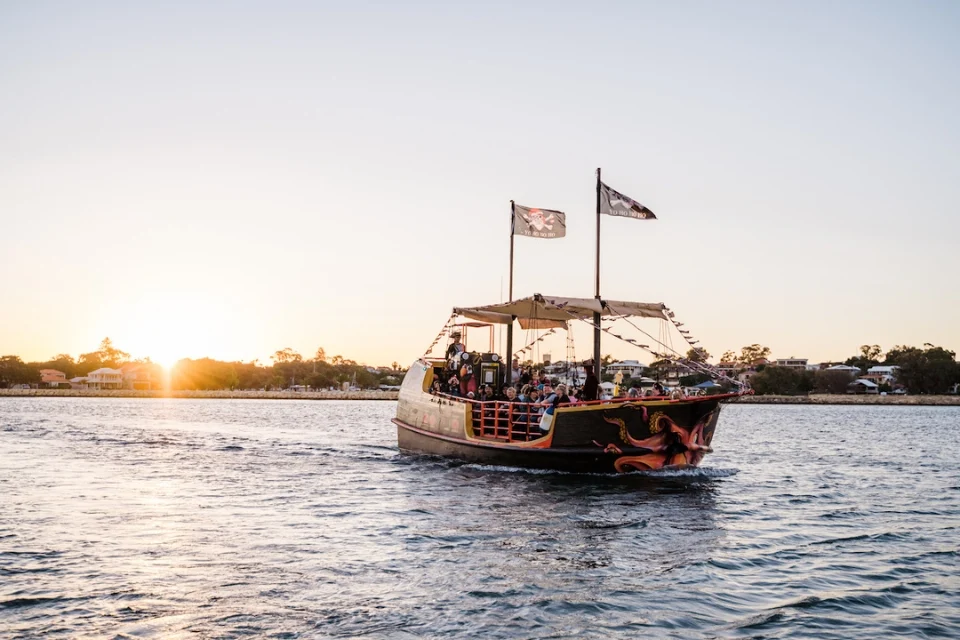 A pirate-themed boat with tattered flags sails on a calm river at sunset. The sky is clear, and the sun is low on the horizon, casting a warm glow over the water. Buildings and trees line the distant shore.