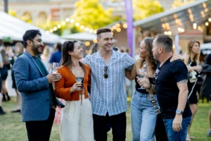 Four people stand outdoors at a festival, smiling and holding wine glasses. A man in the center has his arms around two women. The words Uncorked Mandurah appear above them. Lights and tents are visible in the background.