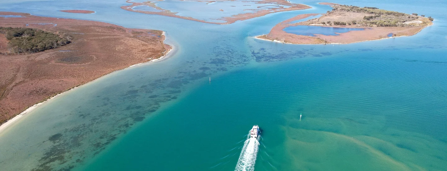 Aerial view of a boat sailing on a clear turquoise calm estuary surrounded by small tree-covered islands and sandbanks. The wake of the boat creates a white trail, and the landscape features contrasting colours of green, blue, and brown.