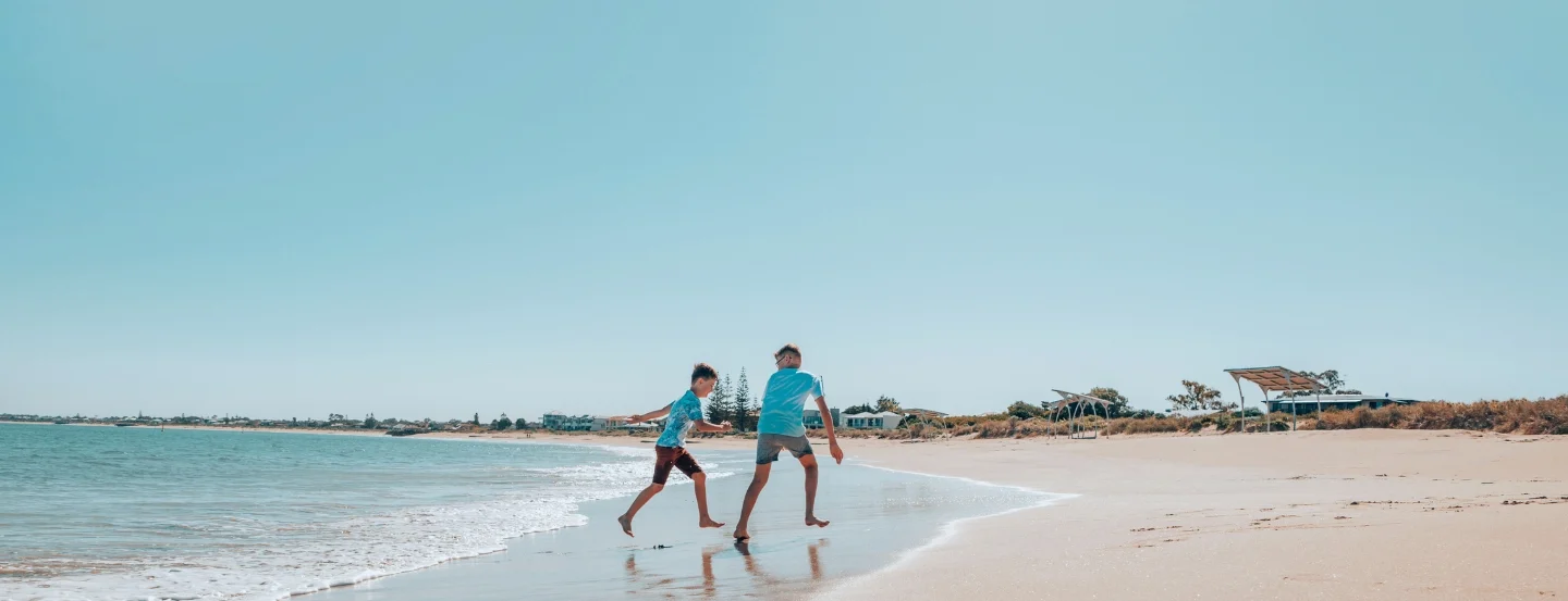 Two boys walk along a sandy beach near the ocean under a clear blue sky. They appear to be wearing shorts and T-shirts. Houses and trees are visible in the distance, lining the coastline.