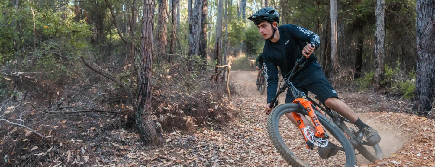 A person wearing a helmet and black gear is riding an orange mountain bike on a dirt trail through a forest. The trail is surrounded by tall trees and scattered leaves, with a sense of motion as the rider leans into a turn.