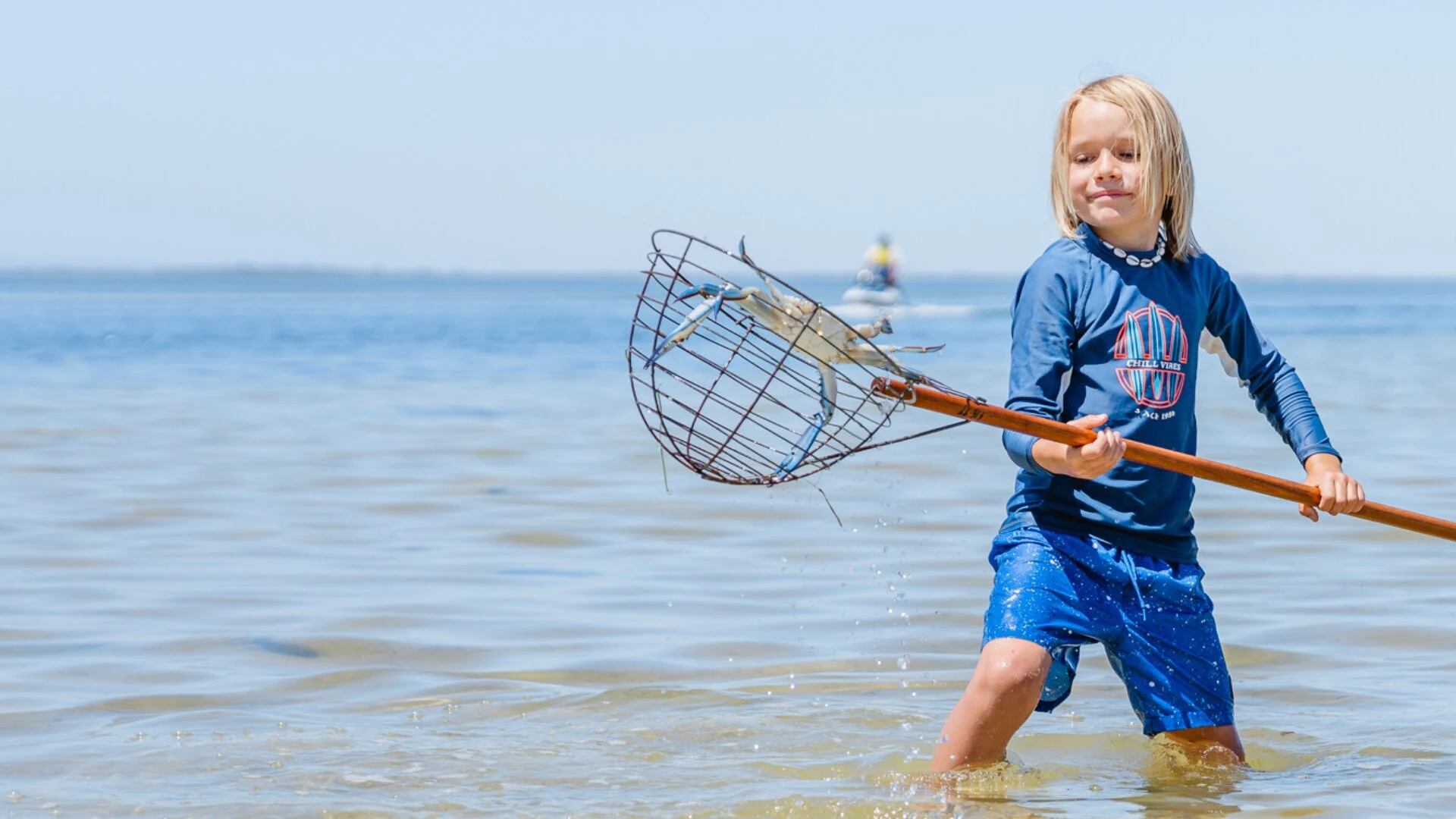 A young blond-haired boy wades through the water, holding a crab scoop.