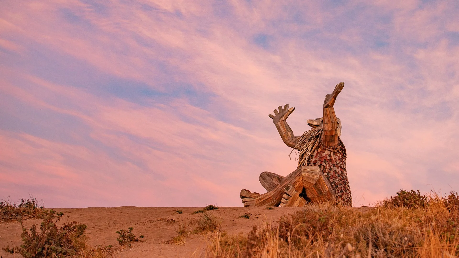 Santi Ikto sculpture, a wooden artwork by Thomas Dambo, at sunset with a pink and orange sky.