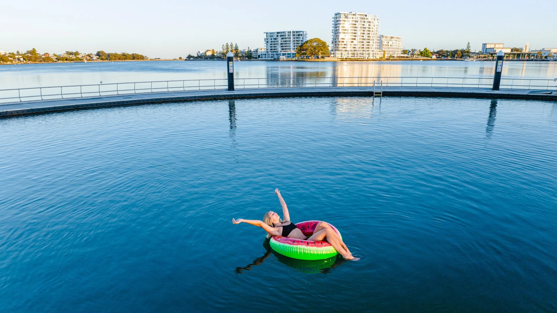 A woman relaxes on a colorful inflatable ring in the middle of Mandurah's Estuary Pool. She is stretching her arms out. In the background, there are buildings and the big figtree along the shoreline under a clear sky.