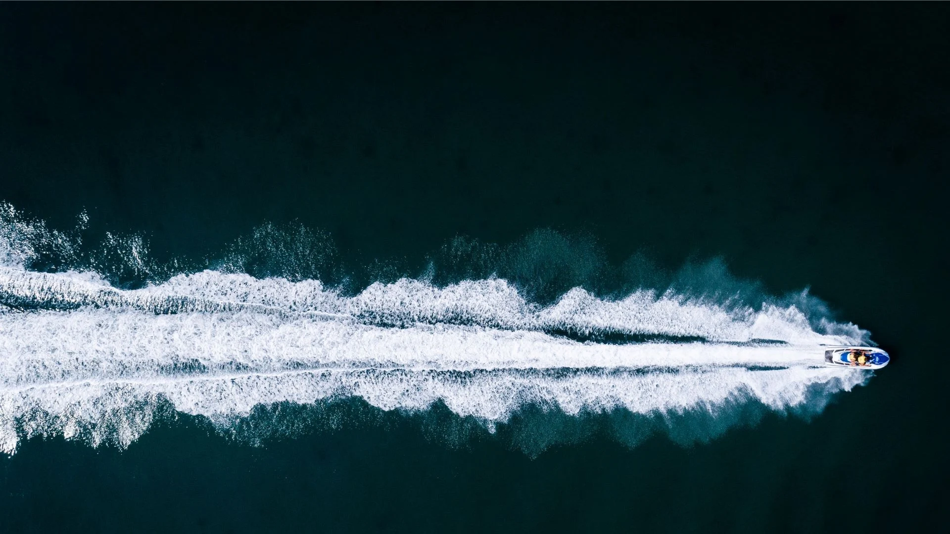 Aerial view of a person on a jet ski speeding across dark water, creating a long, white wake behind. The wake forms a striking contrast against the deep green-blue of the water.