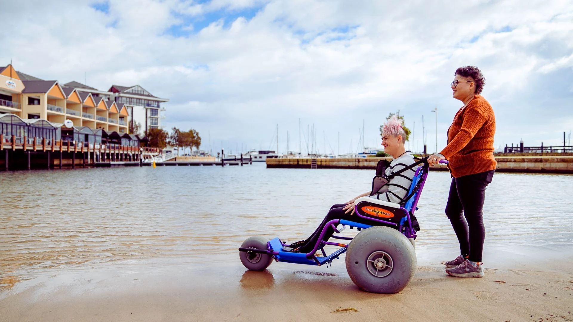 Two women are on a sandy beach near the water. A woman with pink hair sits in a blue beach wheelchair with large wheels, while the other stands behind, holding the handles. Buildings and boats are visible in the background under a cloudy sky.