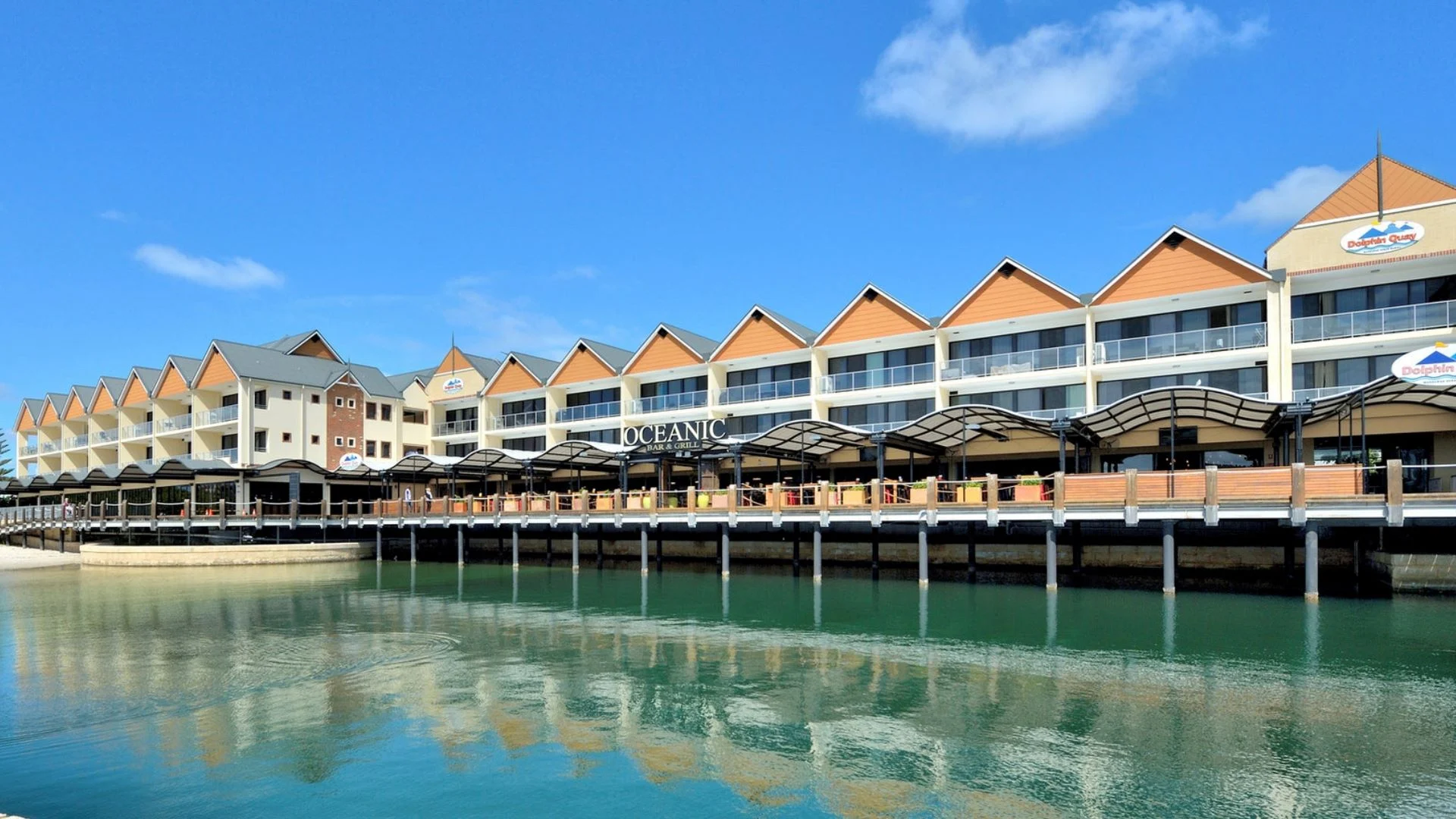A row of modern buildings with peaked roofs along a waterfront. The structures are reflected in the calm water below. The sky is blue with scattered clouds.