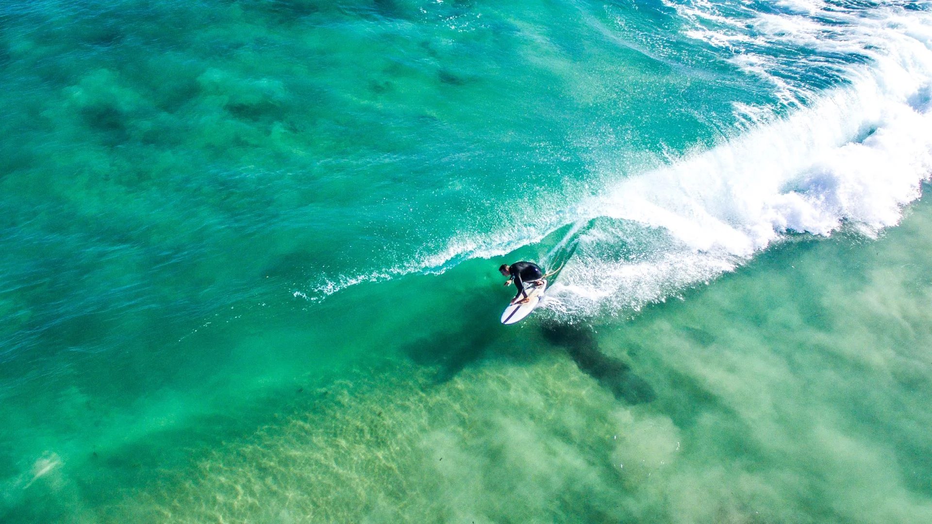 Aerial view of a surfer riding a clear turquoise wave on a sunny day. The surfboard cuts through the water, creating a splash, while the wave curls with white foam. The ocean floor is visible through the transparent water.