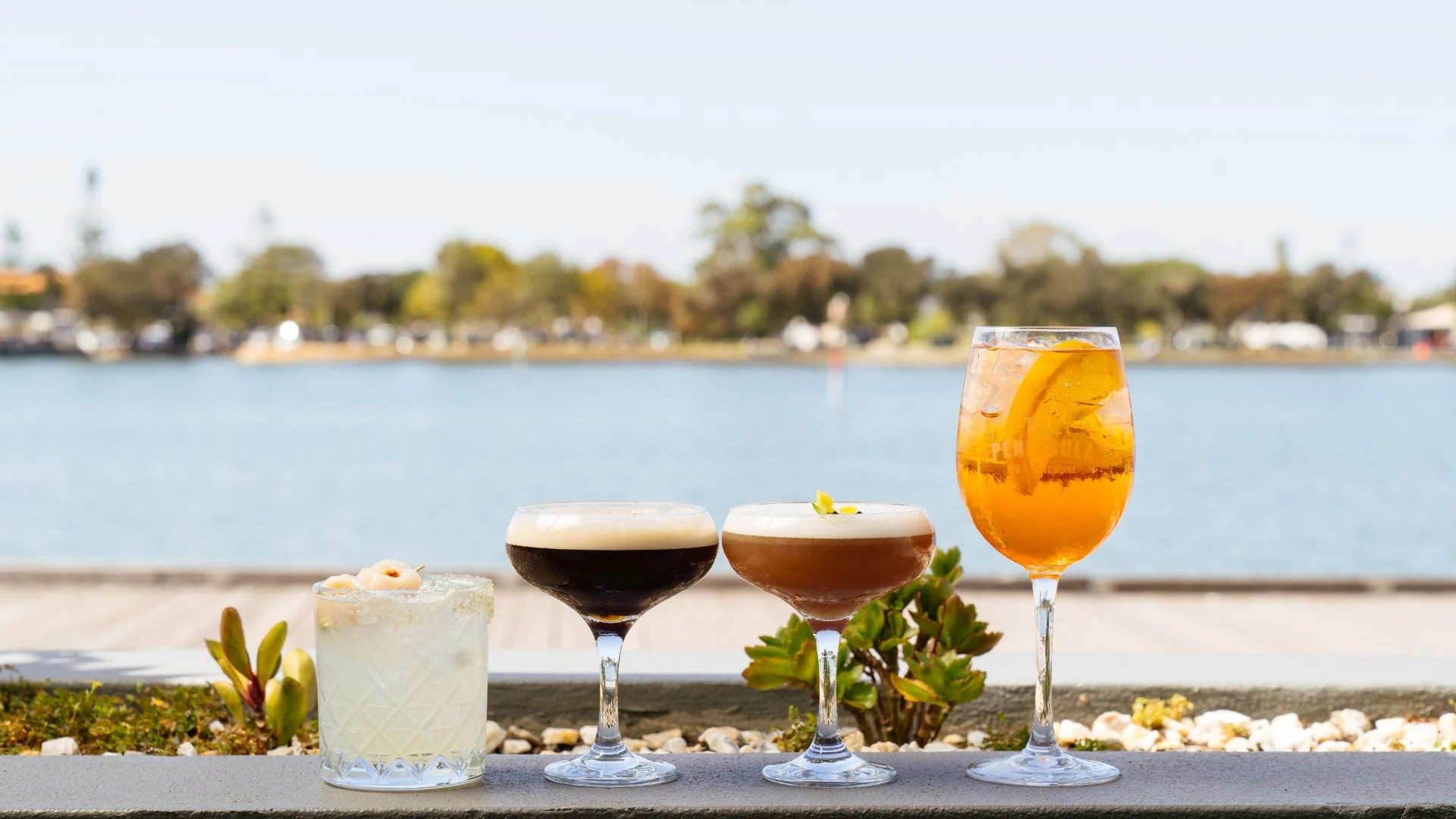Four cocktails are lined up on a stone ledge with a scenic waterfront view in the background. The drinks vary in colour and style, including a light drink with a garnish, two creamy cocktails, and a bright orange beverage with an orange slice.