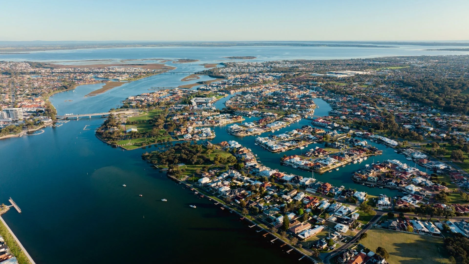 Aerial view of the coastal city Mandurah with winding waterways and residential areas. The landscape features numerous houses, roads, and green spaces, surrounded by expansive bodies of water under a clear blue sky.