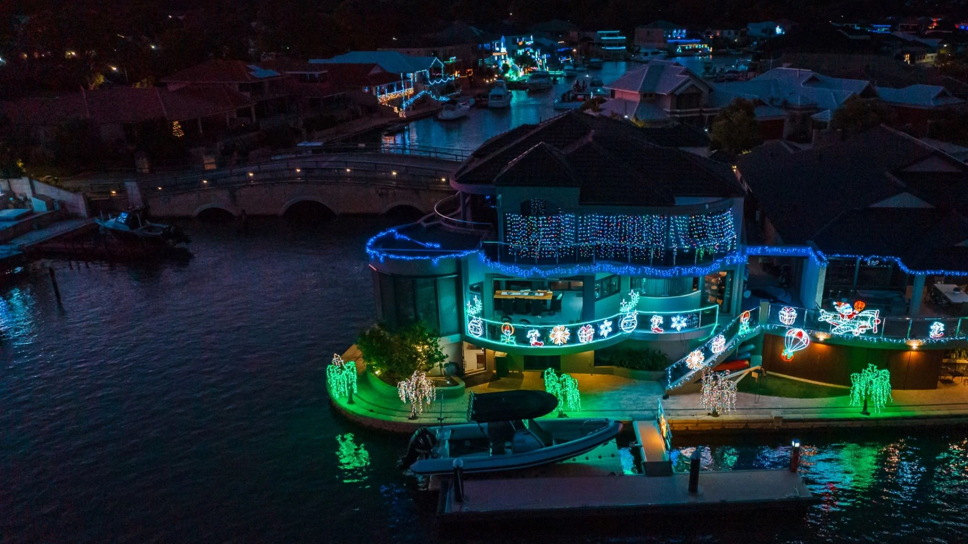 A waterfront house decorated with colorful Christmas lights, including blue and green strings and illuminated ornaments. Theres a boat docked nearby and a lit-up neighborhood visible across the water.