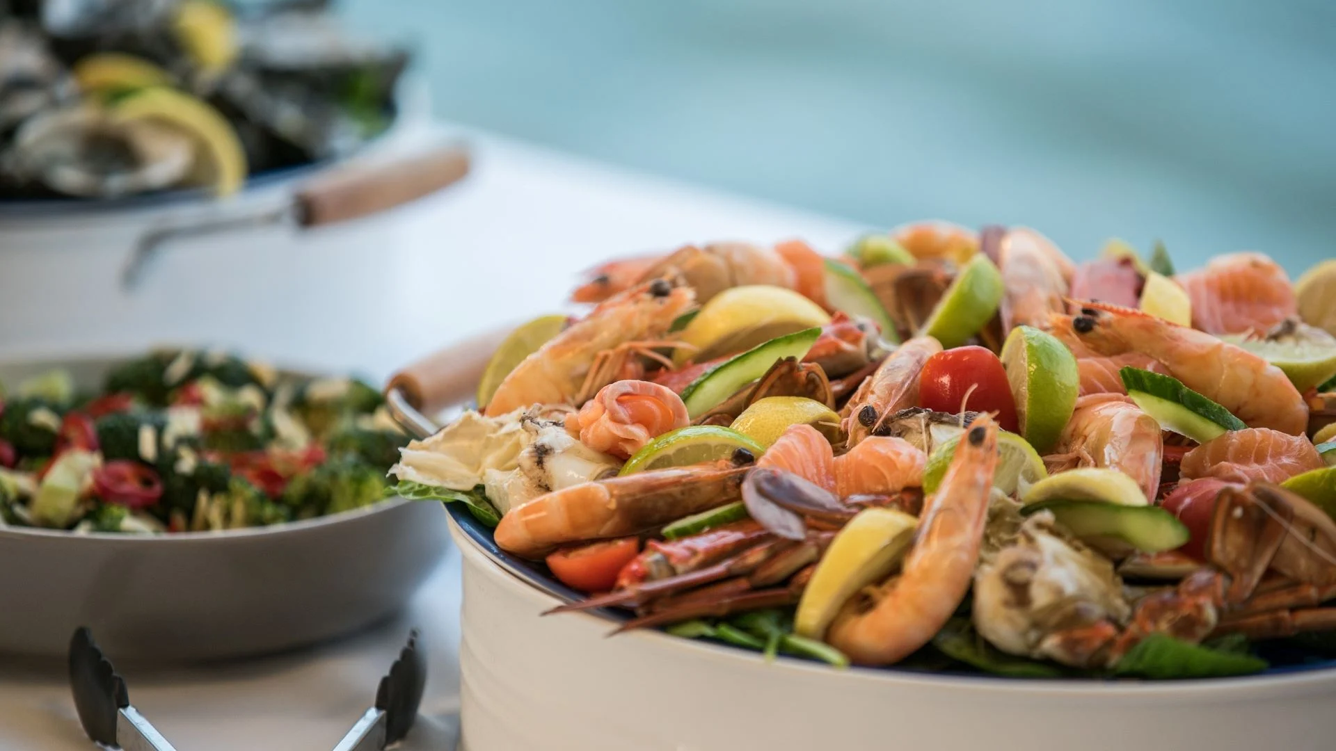 A platter of seafood with shrimp, crab legs, and lemon wedges is displayed prominently. In the background, a salad with greens, cherry tomatoes, and red peppers is visible, along with serving tongs on the side.