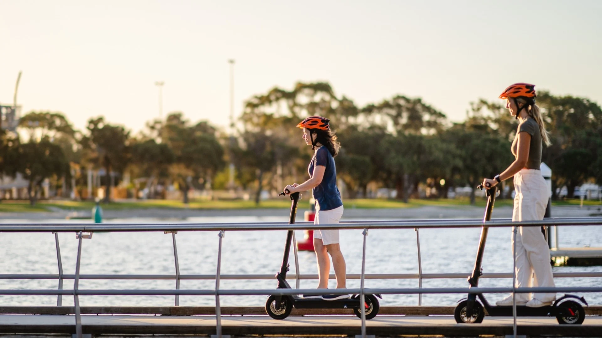 Two people are riding electric scooters on a boardwalk by the estuary at sunset. Both are wearing helmets. Trees and the estuary are in the background.