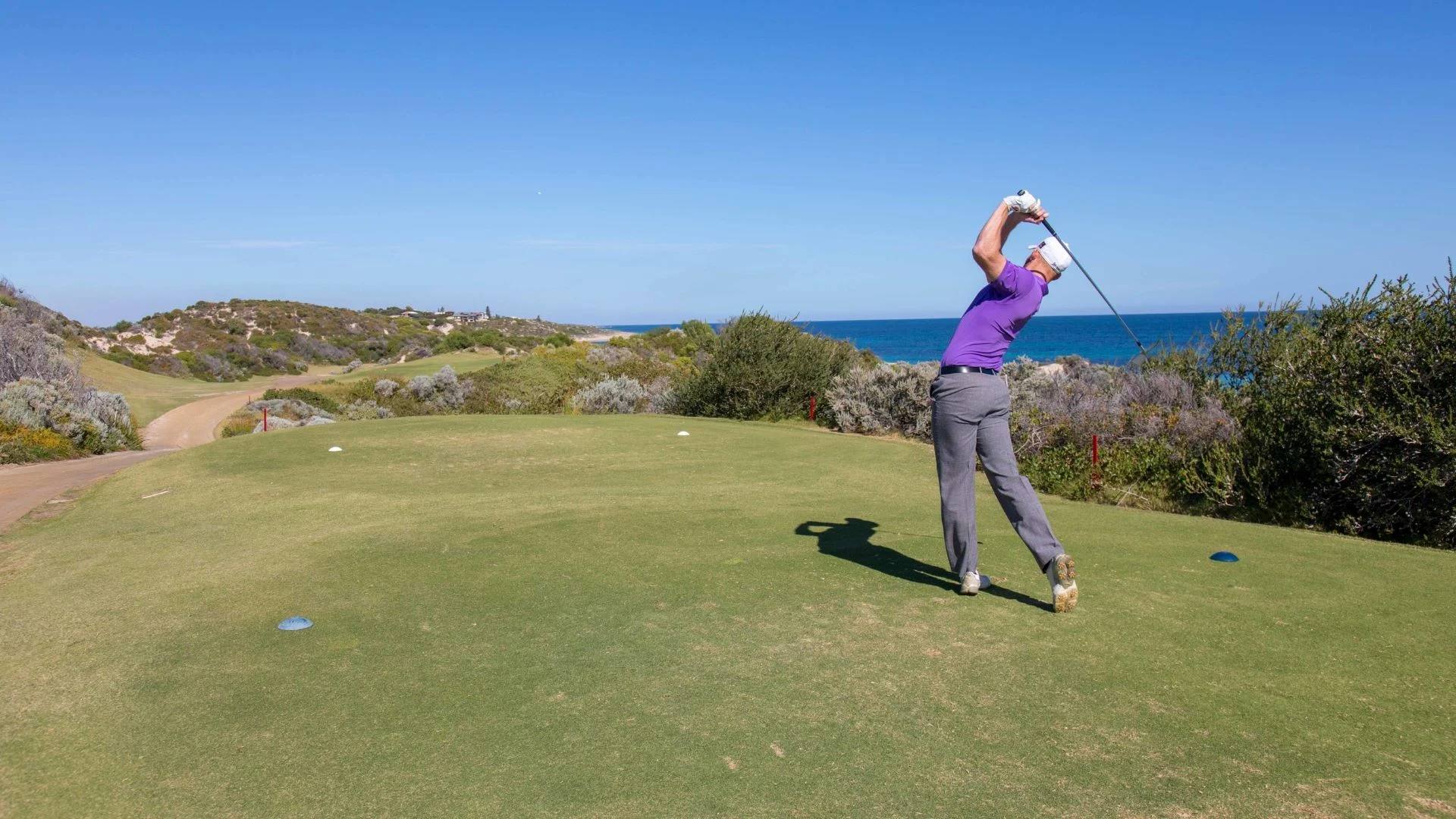 A golfer in a purple shirt and gray pants swings a golf club on a sunny day. The golfer is on a grassy course near a coastline, with a blue ocean and clear sky in the background.