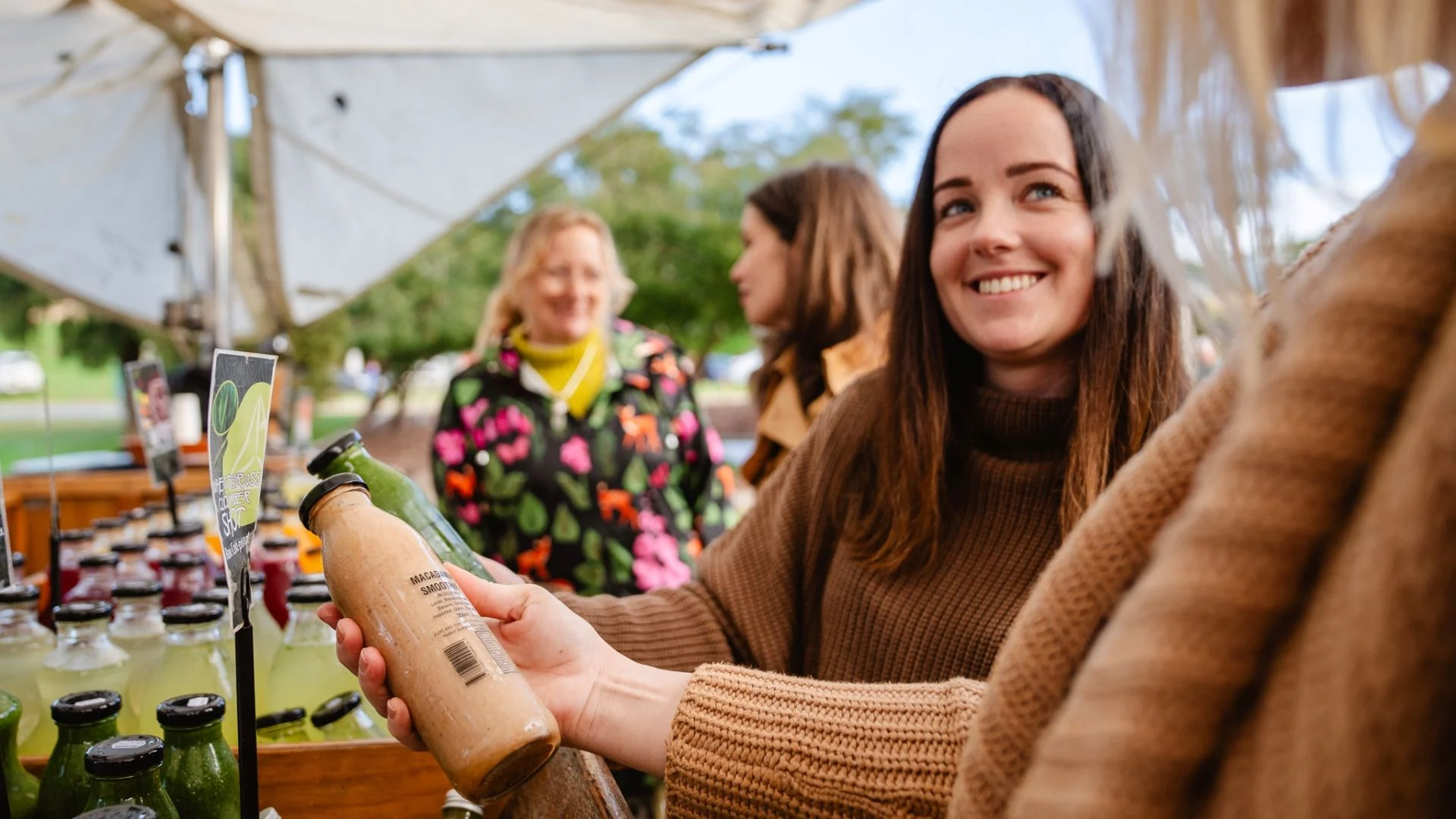 A woman in a brown sweater smiles while holding a bottle of juice at an outdoor market stall. Various juice bottles are displayed on the table. Two other people are in the background, with a sunny day and trees visible.