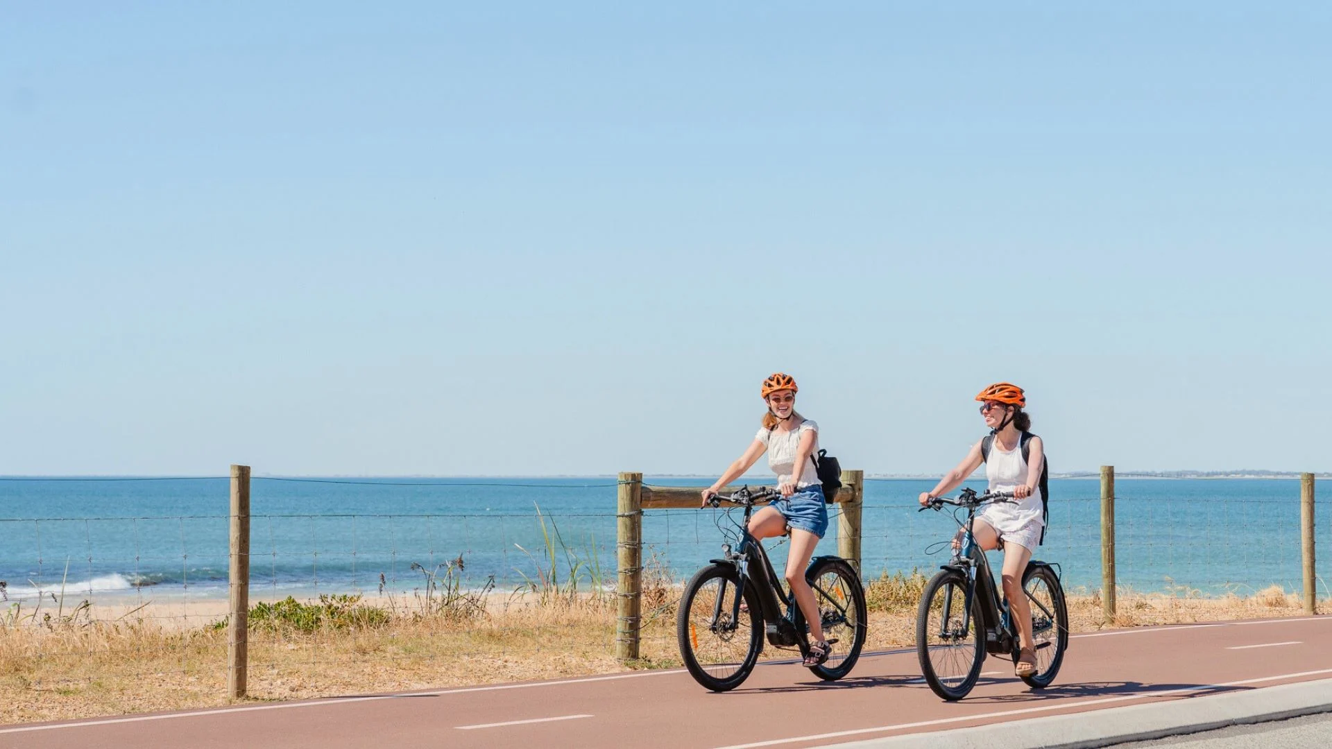 Two women riding bicycles on a coastal path beside a beach. They are wearing orange helmets and casual summer clothes. The sky is clear and the ocean is visible in the background. The path is lined with a simple fence.