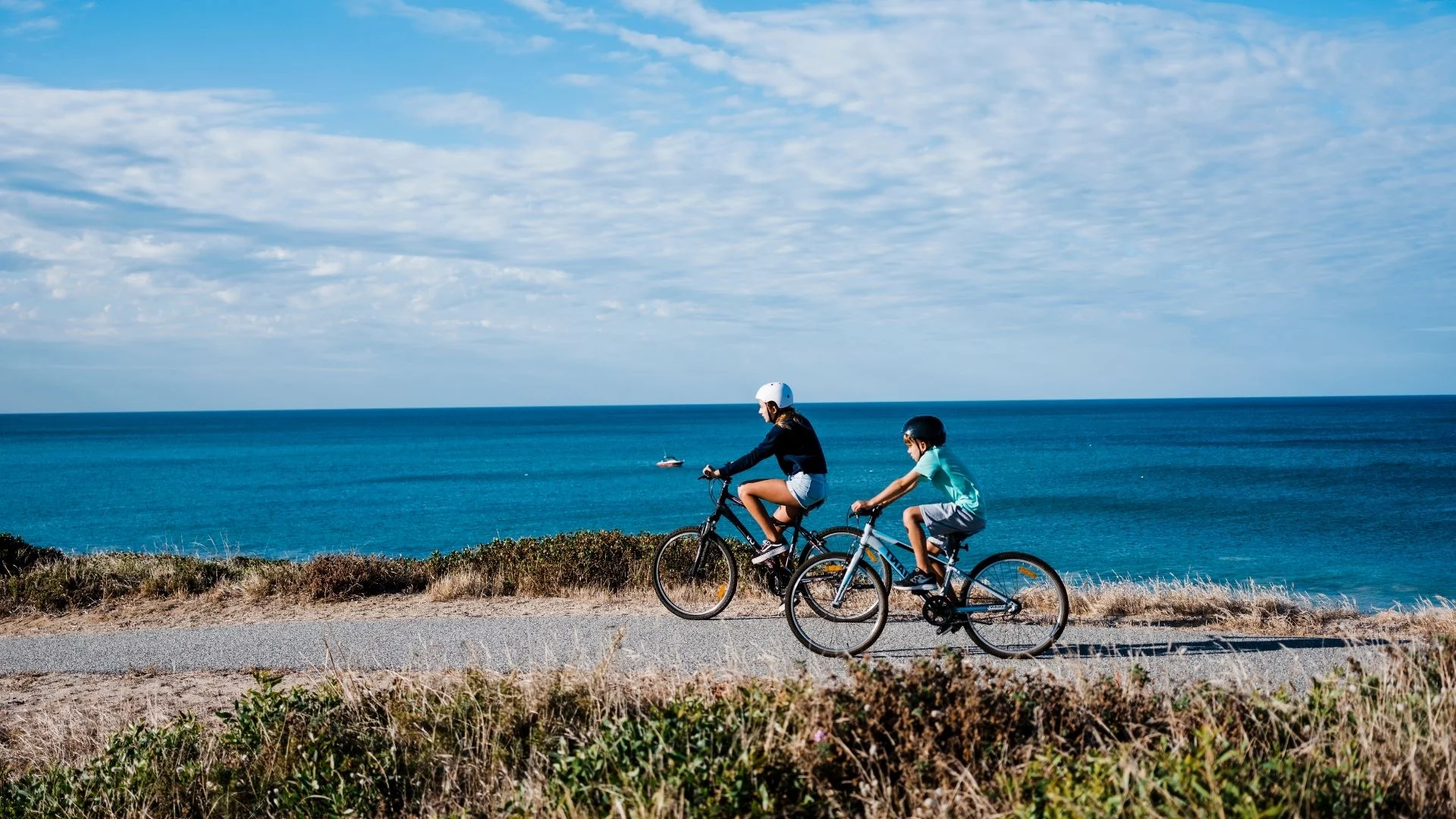 A girl and a boy ride bicycles on a coastal path with the ocean and a partly cloudy sky in the background. The path is surrounded by grass and brush, creating a serene seaside scene.