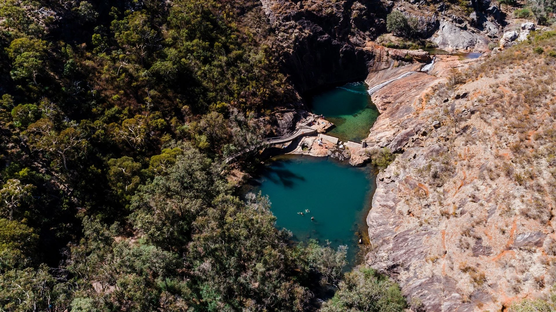Aerial view of Serpentine Falls National Park, a rocky landscape with two natural pools of emerald water surrounded by steep cliffs and dense trees. A narrow footbridge crosses one pool. Swimmers are visible in the water.