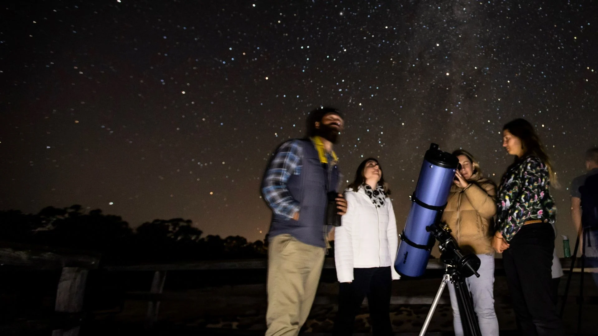 A group of people stargazing at night with a telescope. The sky is clear and filled with numerous stars. They are in a park with silhouettes of trees in the background.