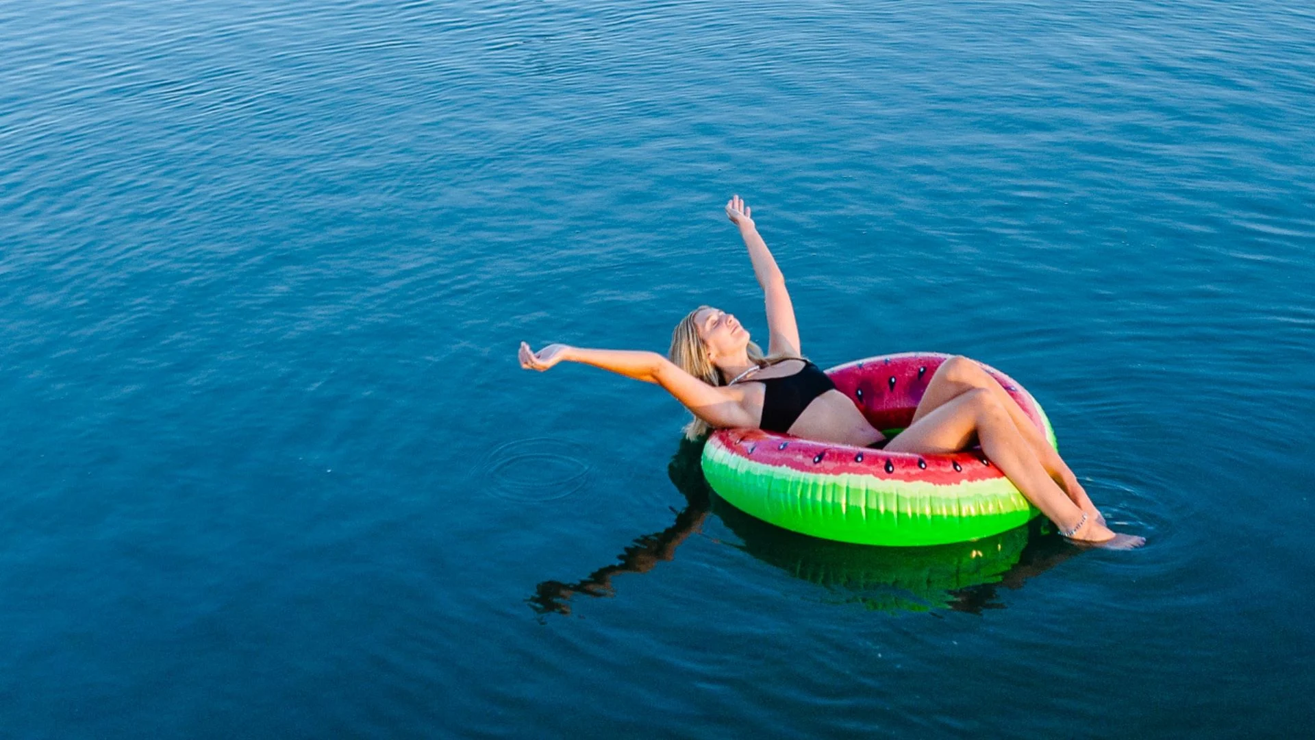 A woman in a black swimsuit relaxes on a watermelon-shaped inflatable float in a calm body of water, with her arms stretched out and eyes closed, enjoying the sun and serene surroundings.
