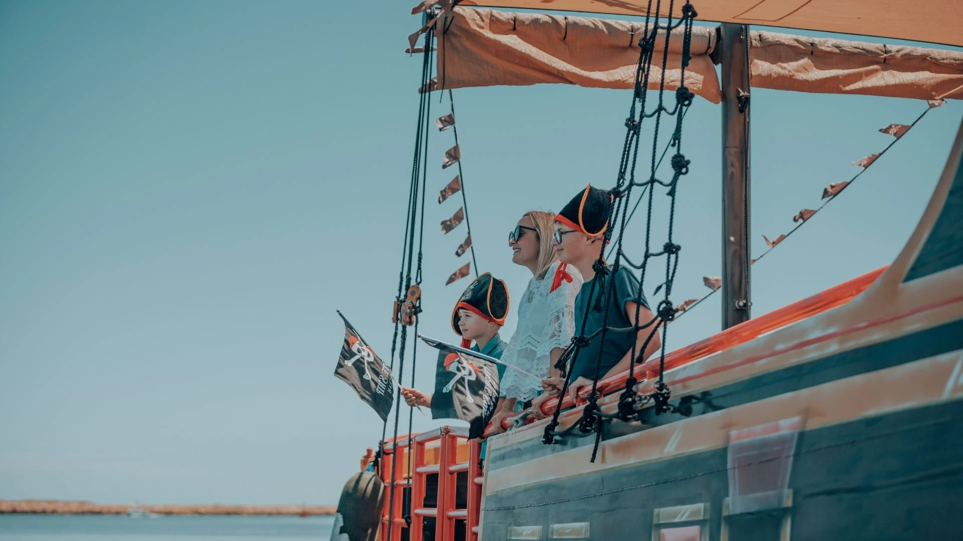 A family dressed in pirate costumes, including hats and bandanas, stands on a wooden ship with a pirate flag. They gaze out at the sea under a clear blue sky.
