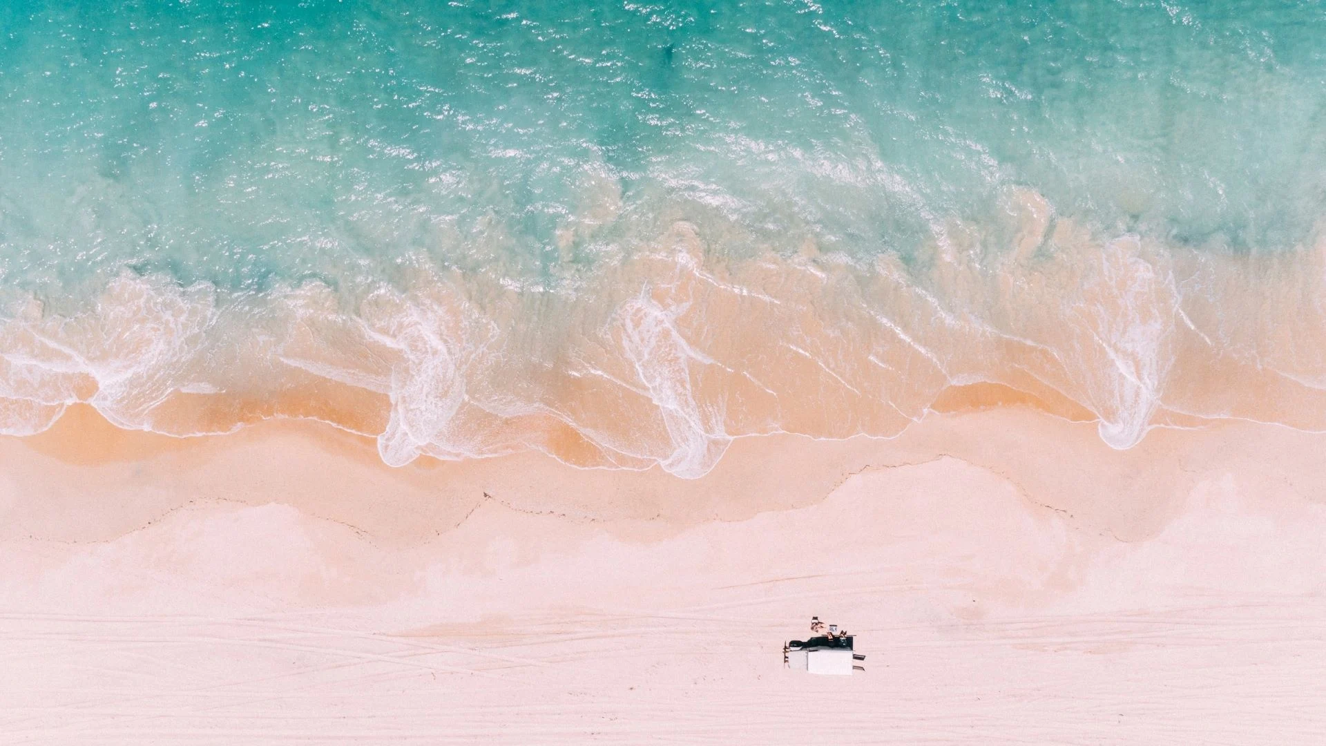 Aerial view of a beach with gentle turquoise waves lapping the sandy shore. A 4WD car is parked on the smooth sand.