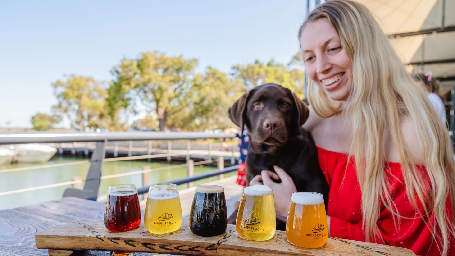 A woman in a red shirt smiles while holding a brown Labrador puppy. In front, theres a wooden paddle with five small glasses of different colored beers. They are sitting outdoors near the estuary with trees in the background.