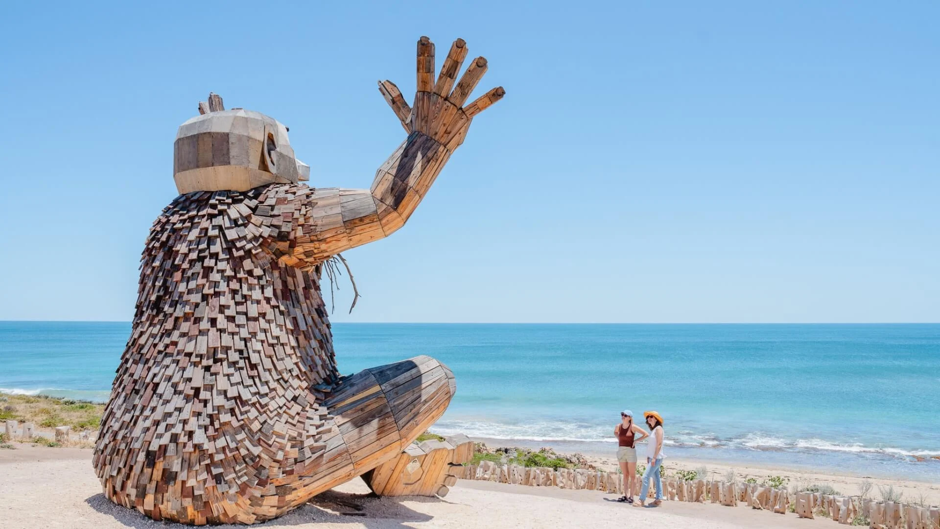 A large wooden sculpture of a Troll figure is seated on the sand, facing the ocean and waving. Two women stand nearby, admiring the artwork. The scene is set against a clear blue sky and calm sea.