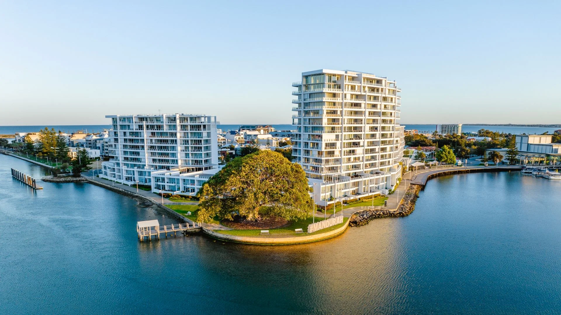 Aerial view of modern waterfront apartment buildings surrounded by Mandjar Bay in Mandurah with a tree-covered island in the foreground. The sunlit scene features clear skies and distant residential and marina areas under a warm evening glow.