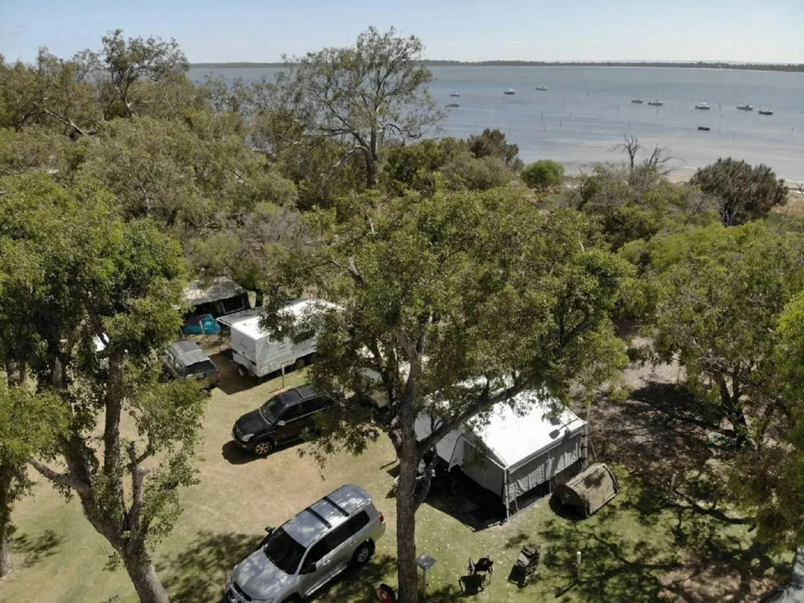 Aerial view of a lakeside campsite with tents, camper trailers, cars, trees, and boats on the water in the background under a clear sky.