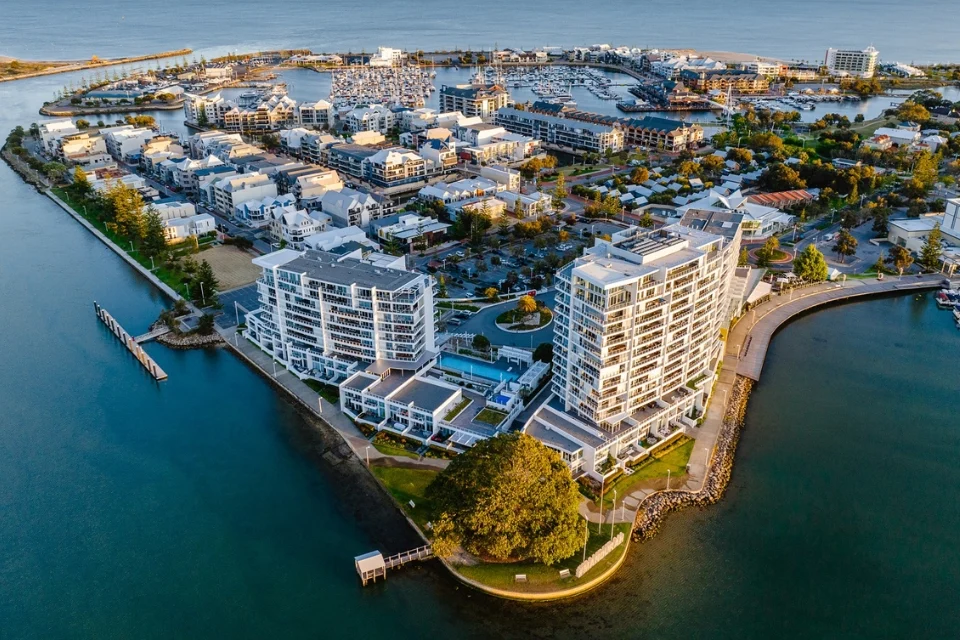 Aerial view of a coastal area with modern buildings and greenery surrounded by water. There are docks, pathways, and residential structures, with the ocean in the background under a clear sky.