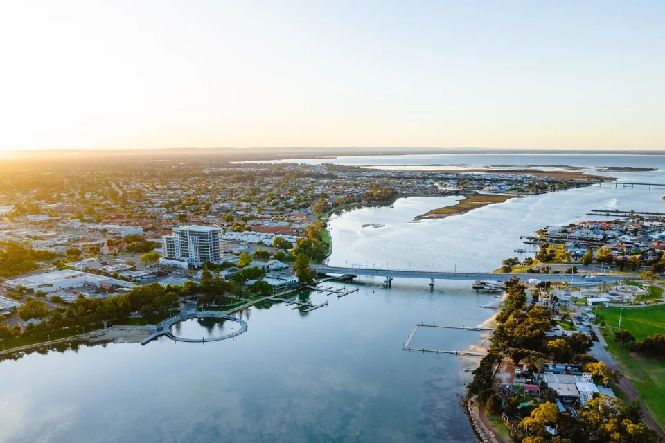 Aerial view of a coastal city at sunset. The image shows a bay with calm waters, a bridge connecting land areas, and a mix of residential and green spaces. The sun casts a warm glow over the scene, creating long shadows.