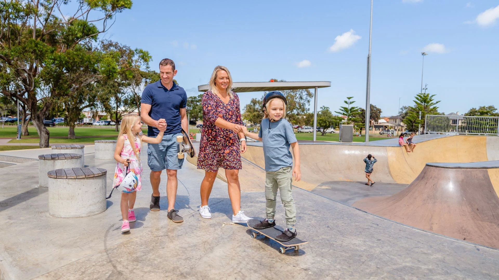 A family enjoys a day at the skate park. A woman guides a young boy on a skateboard, while a man holds a little girls hand. Children play in the background. Trees and a clear blue sky frame the scene.