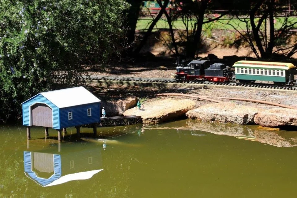 A blue model boathouse is reflected in a pond surrounded by rocks. Nearby, a miniature black and green train with red details travels on a small track. Lush greenery is in the background under bright sunlight.