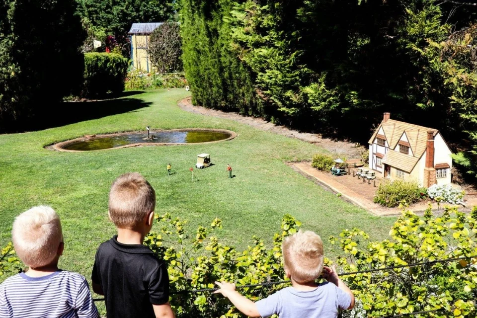 Three children look at a miniature village with small houses and figurines. A pond sits nearby, surrounded by green grass and tall trees. The scene is sunny and peaceful, resembling a tiny world within a garden