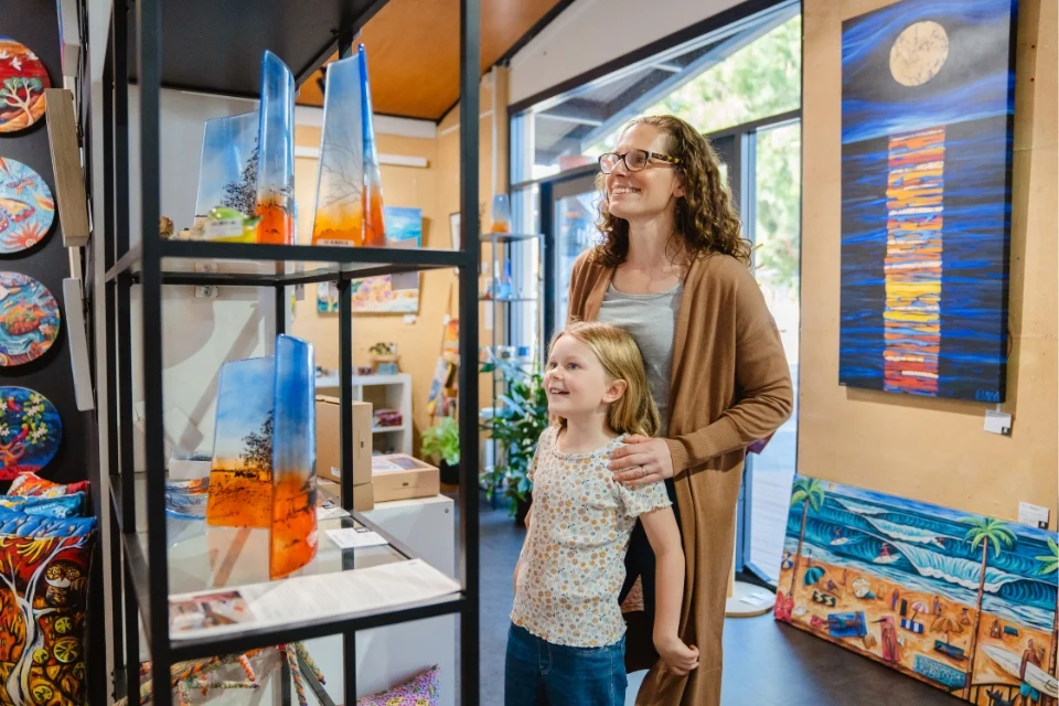 A woman and a young girl, both smiling, admire colourful glass artwork on display in a well-lit gallery. The walls feature vibrant paintings, and a large window lets in natural light, enhancing the bright and creative atmosphere.