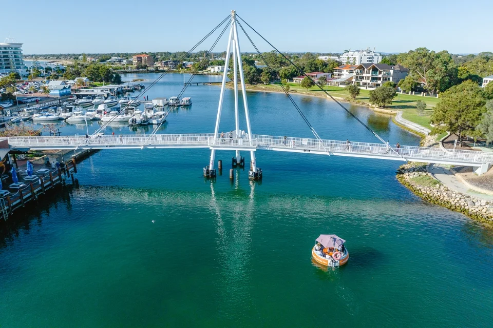 Aerial view of a pedestrian bridge over calm blue water, with a circular boat nearby. Surrounding the waterway are green spaces, trees, and buildings, with a marina full of boats in the background under a clear blue sky.